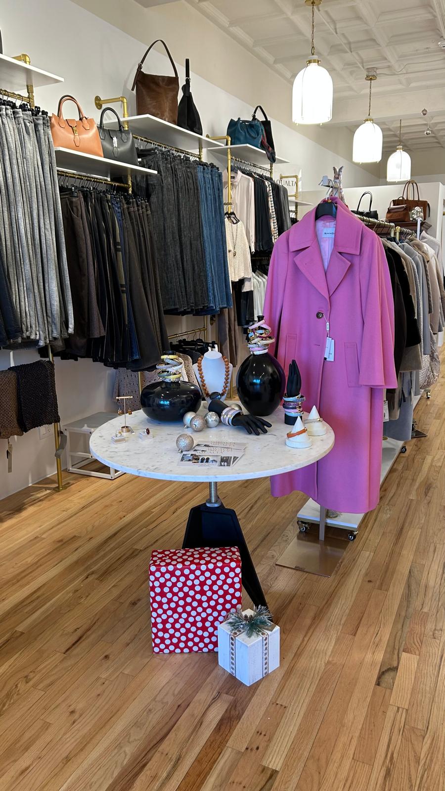 Festive display inside the AqualtaWear boutique featuring a pink wool coat, designer handbags, jewelry, and holiday gift accents arranged on a round table.
