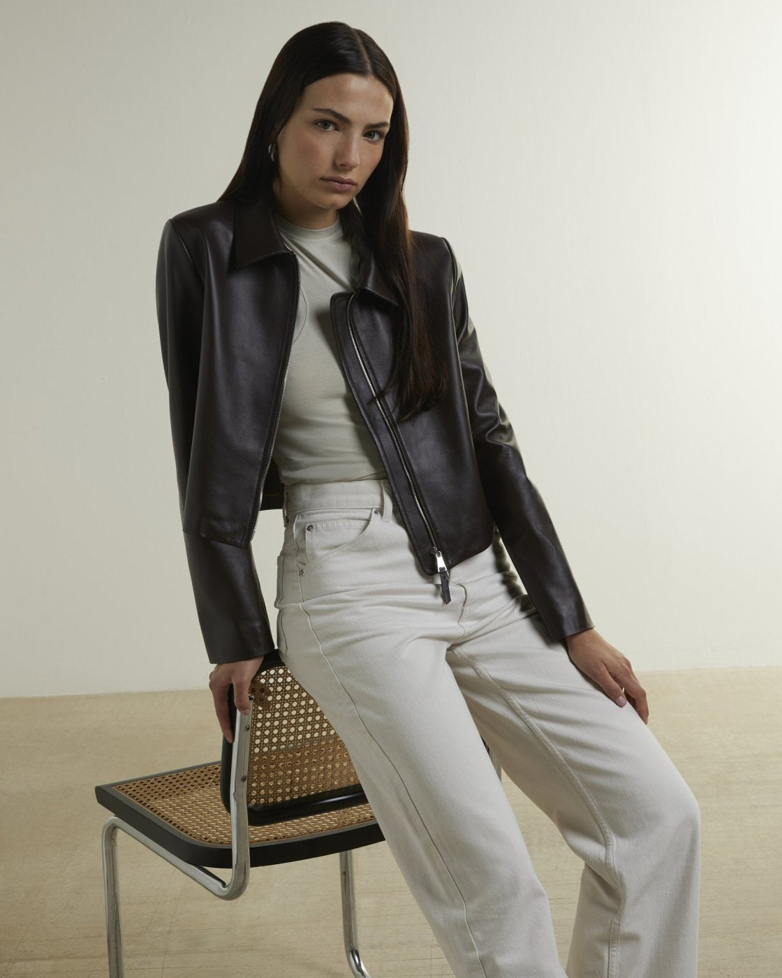 A woman models a black leather moto jacket over a cream ribbed top and cream wide-leg jeans, seated on a modern chair against a minimalist beige studio background.